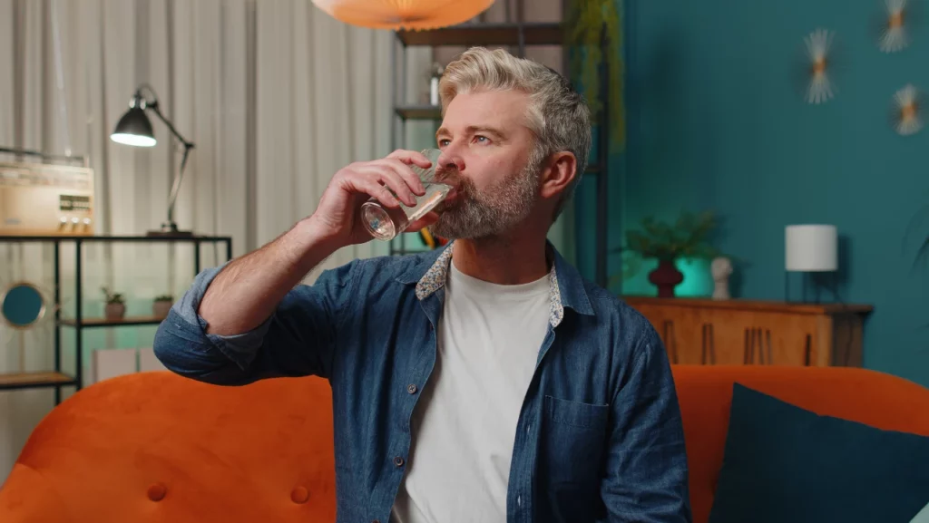 thirsty-middle-aged-man-sitting-at-home-holding-glass-of-natural-aqua-make-sips-drinking-still-water